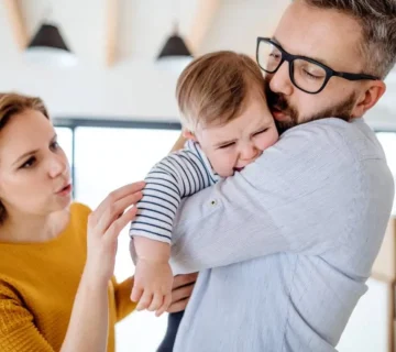 A young family with a crying toddler girl standing indoors at home 360x320